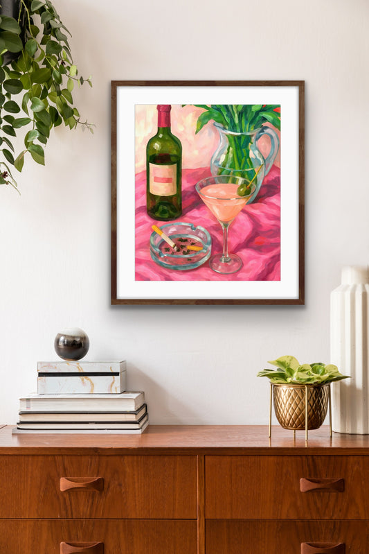 Framed still-life artwork hanging on a light-colored wall above a wooden sideboard with books, decorative objects, and small plants. The artwork depicts a pink tablecloth with a green wine bottle, a cocktail glass with a cherry, and an ashtray with cigarette butts, alongside a glass pitcher holding green leaves. The composition features bright, expressive colors and loose brushstrokes, creating a lively and intimate tabletop scene.