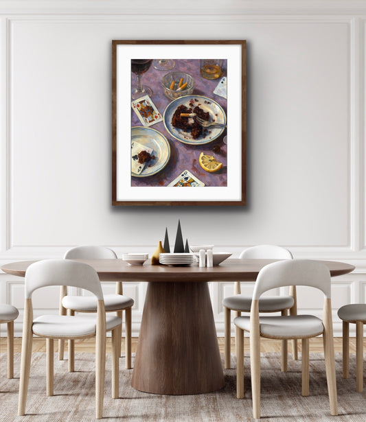 Framed still-life hanging on a white wall above a modern wooden dining table with light upholstered chairs. The artwork depicts a casually arranged table scene with plates of chocolate cake, a fork, a glass of red wine, an ashtray with cigarette butts, a lemon wedge, and scattered playing cards on a purple tablecloth. The artwork has warm tones and expressive brushstrokes, creating a nostalgic, intimate atmosphere.