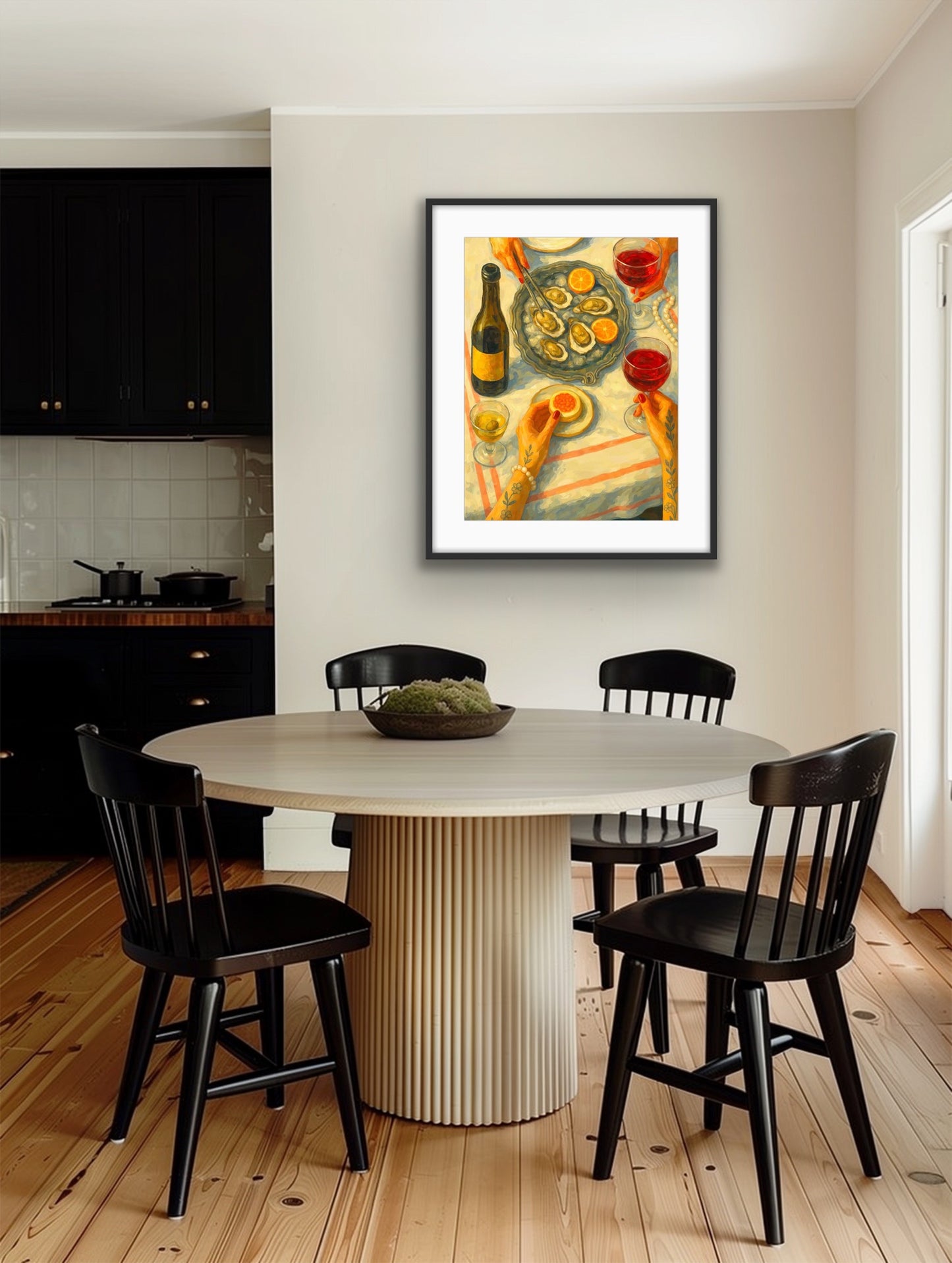 Framed still-life artwork hanging on a light wall above a round dining table with black wooden chairs in a modern kitchen-dining space. The artwork shows a tabletop scene with oysters on a platter, lemon slices, glasses of red and white wine, and a bottle, with two tattooed hands reaching into the frame. Painted in warm tones with visible brushstrokes, the composition captures a lively shared meal from a top-down perspective.